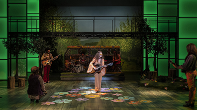 On a stage of green lighted panels framing a colorful VW bus amid trees and on a stage light with flower patterns stands Dame Senior playing guitar with other members of the band on the periphery. 